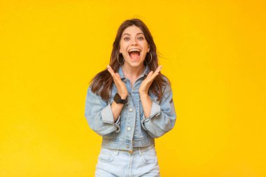 Portrait of surprised beautiful brunette woman with makeup in denim casual style standing and looking at camera with open mouth and amazed face on yellow background clipart