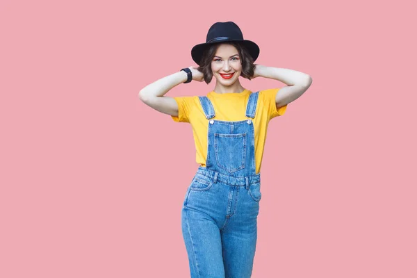 シャツと青デニムのオーバー オール メイクと黒い帽子調達の腕のポーズとピンクの背景に こぼれるような笑顔でカメラ目線の幸せな女性の肖像画 — ストック写真