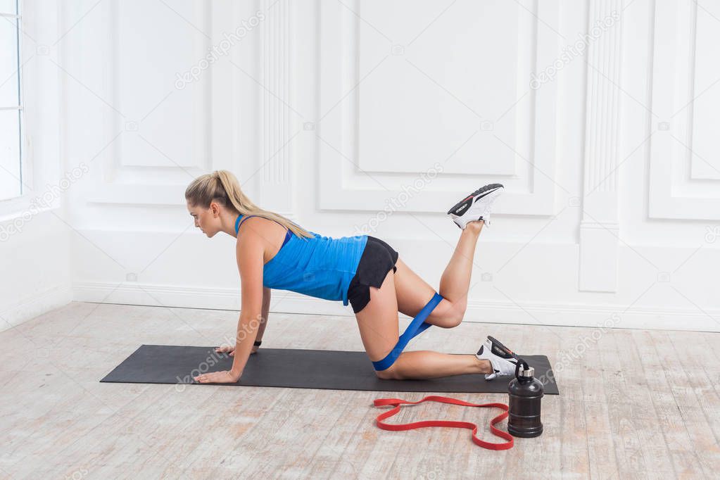 Side view of strong and fit athletic blonde woman in black shorts and blue top with bands training legs and glutes muscular with expander, sport and healthy concept 
