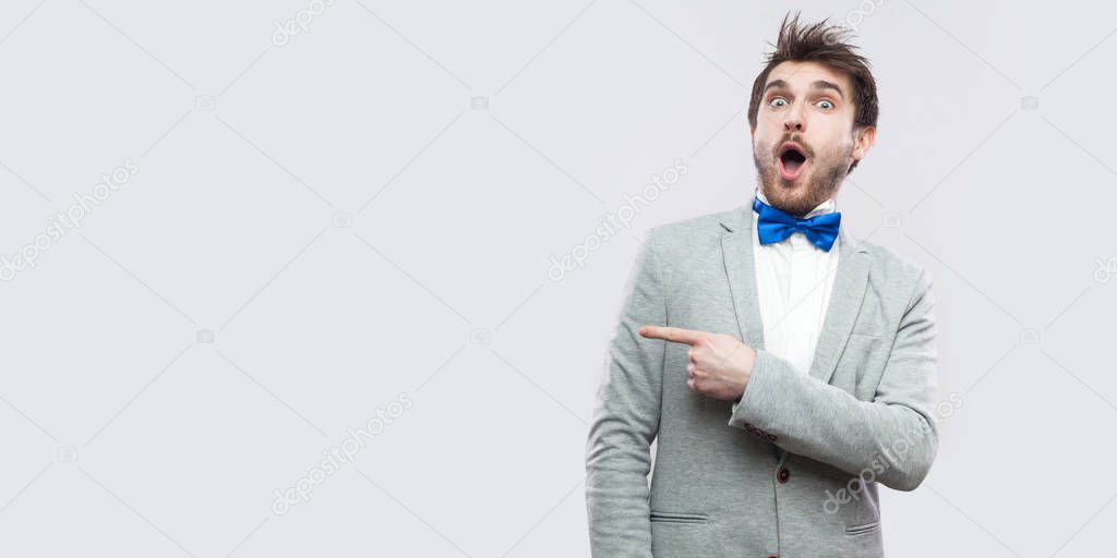 Portrait of amazed handsome bearded man in casual grey suit and blue bow tie standing and looking at camera with surprised face while  pointing aside on light grey background.