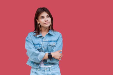 Portrait of thoughtful beautiful brunette young woman with makeup in denim casual style standing with crossed hands and looking away on red background. clipart