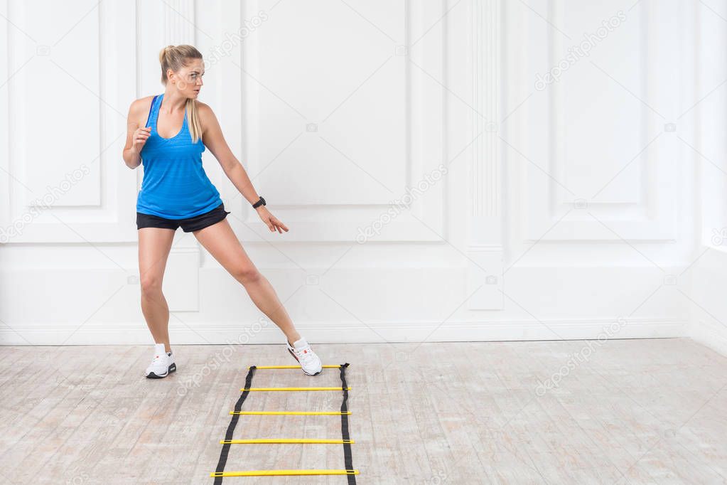 Full length of sporty beautiful young athletic blonde woman in black shorts and blue top doing cardio workout and training on agility ladder drills on floor in gym