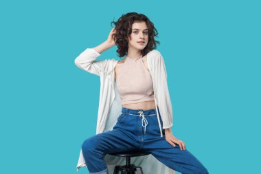 Sandalye üzerinde oturan ve mavi arka planda gülümseme ile kamera bakarken moda modeli gibi poz Casual tarzı memnun güzel genç esmer kadın portre