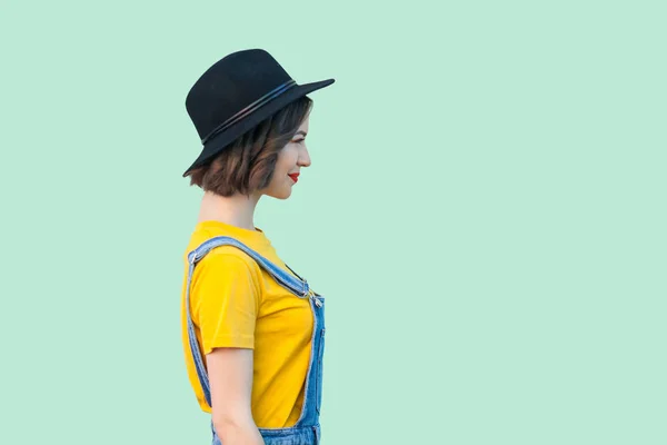 青いデニムのオーバー オールと笑顔と明るい緑の背景にまっすぐ見ている黒い帽子の黄色の シャツでかなり若い流行に敏感な女性の側面図 — ストック写真