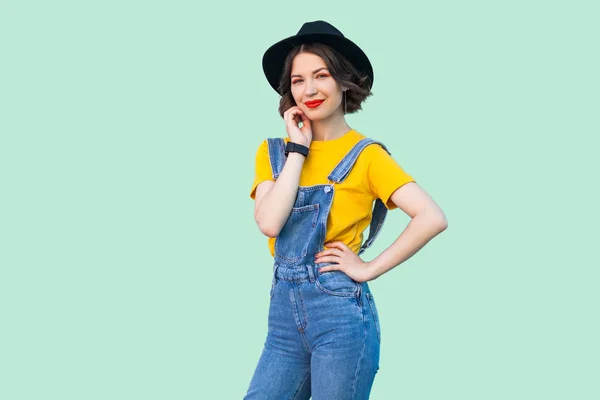 青いデニムのオーバー オールと黒い帽子立っている腰に手と明るい緑の背景にカメラ目線と黄色の シャツでかなり若い流行に敏感な女性の肖像画 — ストック写真