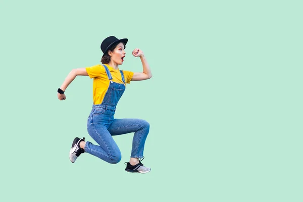side view of surprised funny hipster woman in blue denim overalls and yellow t-shirt with black hat jumping in super mario style on light green background