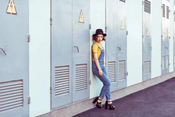 身穿蓝色粗斜纹棉布工作服和黄色 T恤的年轻女子在工业建筑附近摆姿势时 穿着黑色帽子 感官看着相机 — 图库照片