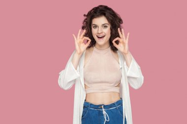 eğlenceli heyecanlı güzel esmer genç kadın kıvırcık saç modeli ile rahat tarzı gösteren ok işaretleri ve kamera pembe arka planda bakıyor.