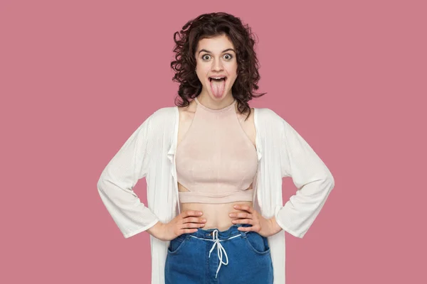 カジュアルなスタイルでカーリー髪型を持つ面白い狂気の美しいブルネットの若い女性は腰に手を保持し ピンクの背景に舌を示します — ストック写真