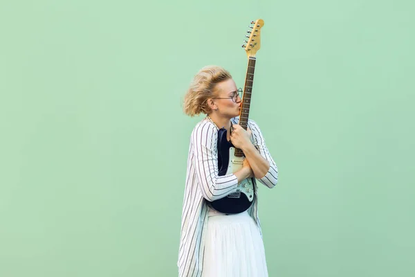 young blonde woman in white clothes and striped blouse with eyeglasses embracing and kissing her lovely electric guitar on light green background.