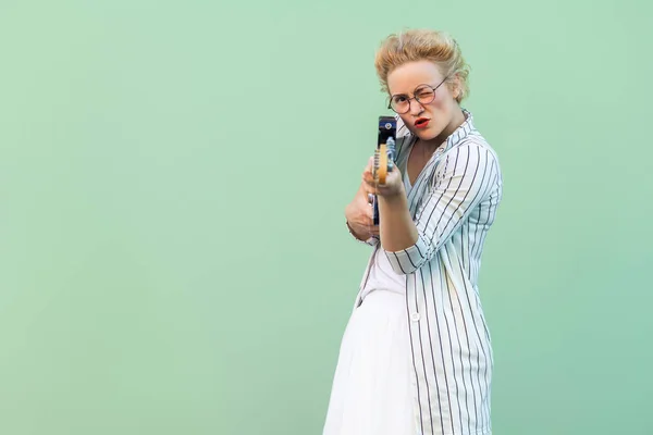 young blonde woman in white clothes and striped blouse with eyeglasses standing holding electric guitar like gun and focusing on camera over light green background.