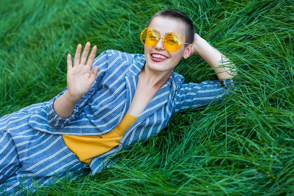 beautiful young woman in casual blue striped suit and yellow shirt with sunglasses lying down on green grass and looking at camera with toothy smile while greeting