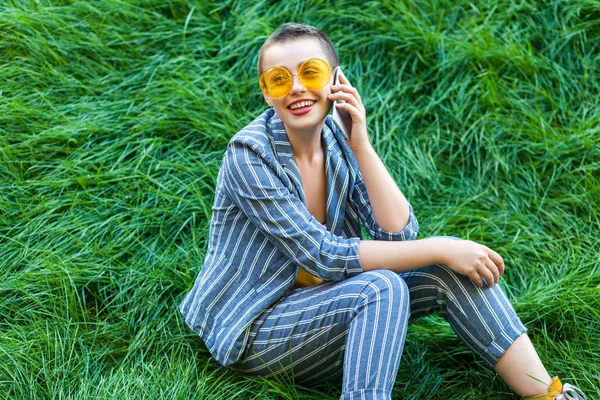 beautiful young woman in casual blue striped suit and yellow shirt with sunglasses sitting on green grass on talking on smartphone and toothy smiling