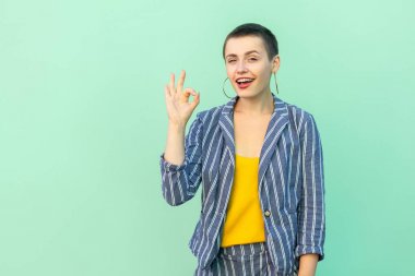 funny beautiful short hair young stylish woman in casual striped suit showing Ok sign and looking at camera with toothy smile on green background  clipart