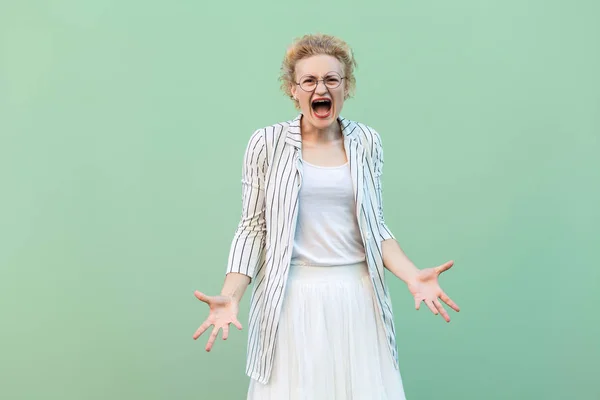 angry young blonde woman in white striped blouse with eyeglasses standing with raised arms and screaming on green background