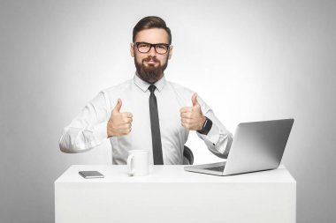 beyaz gömlek ve siyah kravat yakışıklı memnun sakallı genç işadamı gülümseyerek ve kameraya bakarken başparmak gösteren ve dizüstü bilgisayar ile ofis masasında otururken 