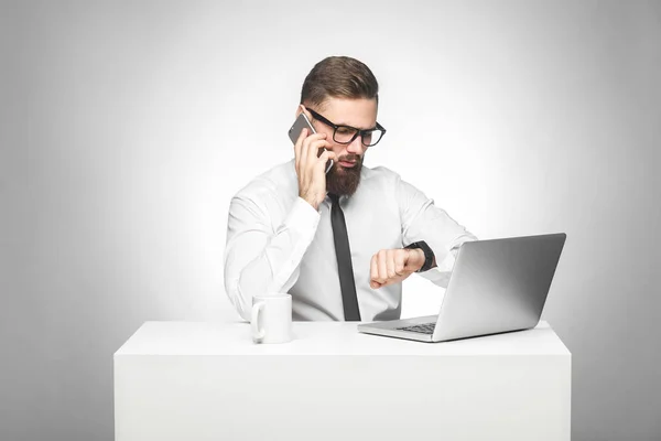 穿着白衬衫和黑色领带的留胡子的年轻工人在智能手机上交谈 在手表上检查时间 而坐在办公室桌前与笔记本电脑 — 图库照片