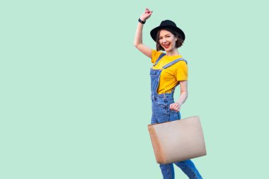 Hipster denim tulum ve siyah şapka yükseltilmiş kol ile mağaza çantası tutan neşeli güzel genç kadın ve yeşil arka plan üzerinde kamera bakıyor