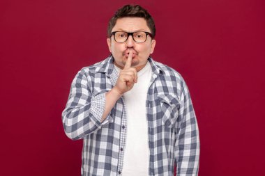 rahat damalı gömlek ve gözlük içinde ciddi yakışıklı orta yaşlı işadamı sessizlik işareti gösteren ve kırmızı arka planda kamera bakarak