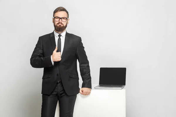 職場の近くに立ち 灰色の背景に親指を表示するインテリジェンス黒いスーツでハンサムな成功したひげの若いビジネスマン — ストック写真