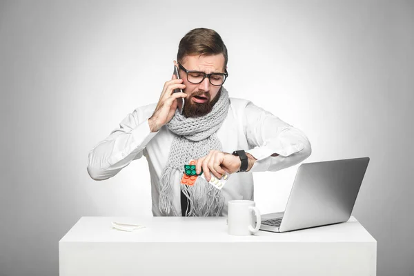 気のついた寒い病気の若い男は 机に座って スマートフォンで話しているときに手に時間をチェックしながら 寒さを病んでいる若者時計 会議のコンセプトを計画 — ストック写真