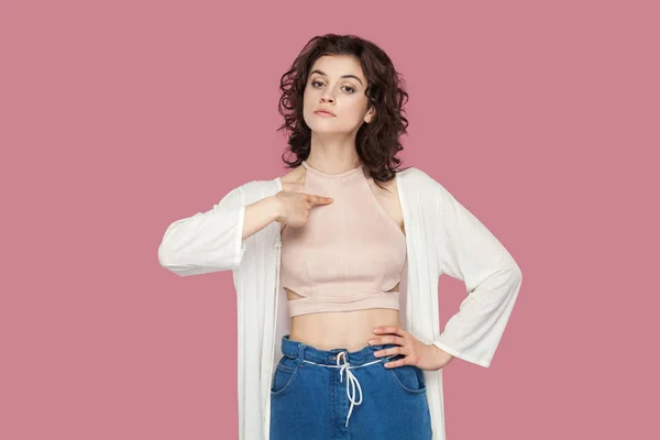 haughty proud beautiful brunette young woman with curly hairstyle in casual style pointing herself and looking at camera on pink background