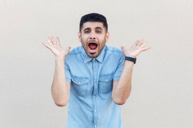 şok yakışıklı genç sakallı adam mavi gömlek ayakta ve açık bej duvar arka plan üzerinde kolları ve sürpriz yüzü ile kamera bakıyor