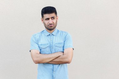 düşünceli karışık yakışıklı genç sakallı adam mavi gömlek çapraz kolları ile ayakta ve uzak hafif bej duvar arka plan bakıyor
