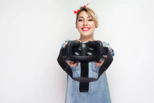 beautiful young woman in casual blue denim shirt and red headband showing virtual reality headset isolated on white background.