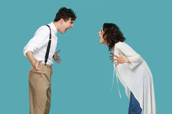 side view of two aggressive brunette partners standing and screaming at each other on blue background, conflict  concept