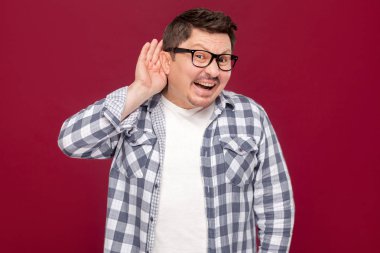komik yakışıklı orta yaşlı işadamı rahat damalı gömlek ve gözlük kulak elle ayakta ve koyu kırmızı arka planda gülümseyerek kameraya bakarak.