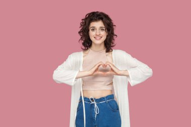 sevgi el jesti ile ayakta ve pembe arka plan üzerinde dişli gülümseme ile kamera bakarak rahat tarzı kıvırcık saç modeli ile güzel esmer genç kadın