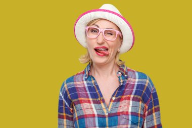 şapka ve gözlük ile rahat tarzı komik deli modern şık olgun kadın dil gösteren ve sarı arka planda kamera bakarken göz kırpma