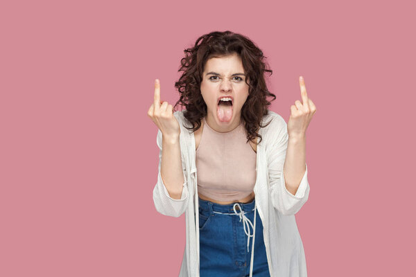 angry screaming brunette young woman with curly hairstyle in casual style looking at camera and showing middle finger fuck sings on pink background