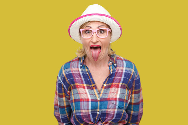  Смешно удивлен современный стильный зрелая женщина в случайном стиле с шляпой и очки показывая язык и глядя на камеру с большими глазами на желтом фоне

