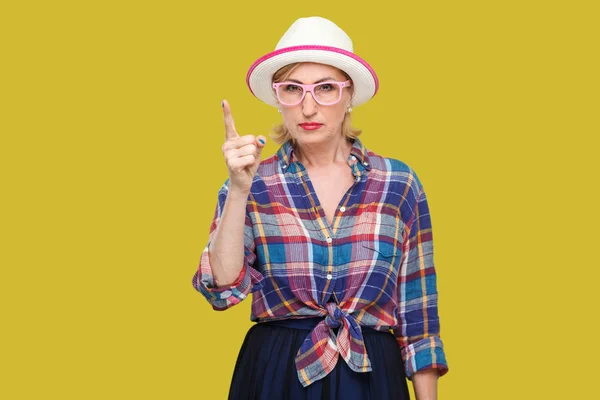严重霸道的现代时尚成熟女人在休闲风格与帽子和眼镜显示警告标志和看相机在黄色背景 — 图库照片