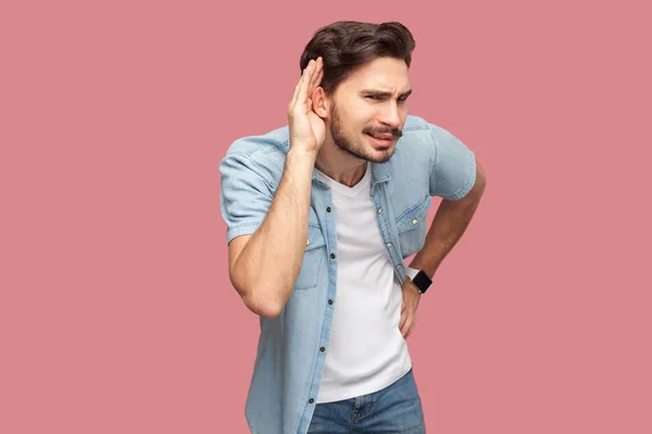 attentive handsome bearded young man in blue casual style shirt standing with hand on ear and trying to hear with serious face on pink background