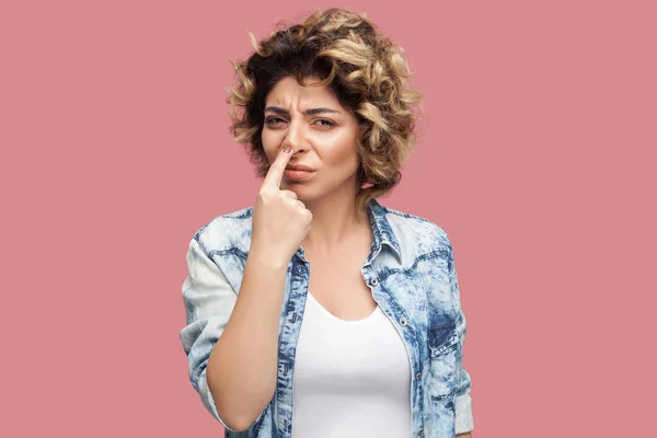 serious young woman with curly hairstyle in casual blue shirt standing with finger on nose and showing lie gesture on pink background
