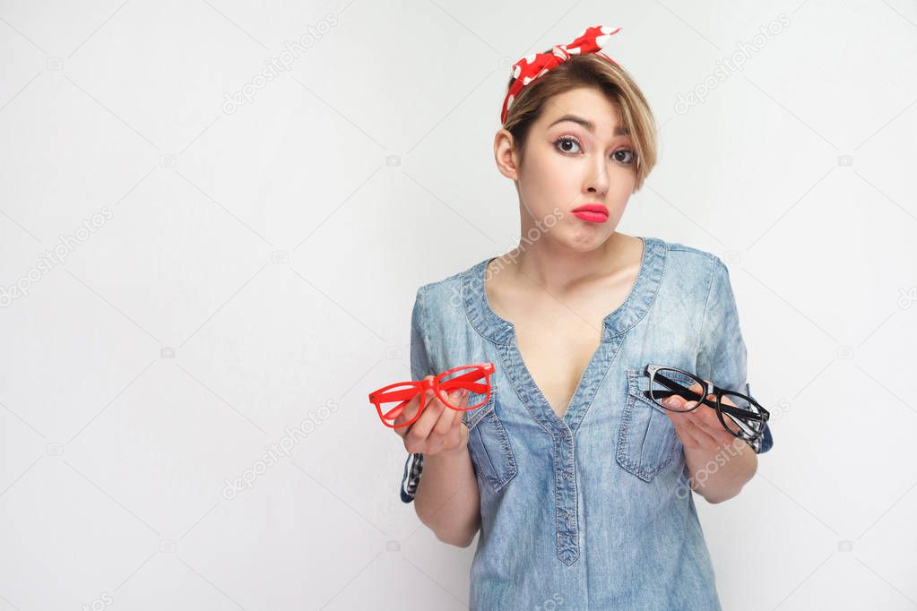 sadness young woman in casual blue denim shirt with makeup and red headband holding two pairs of glasses with doubt face isolated on white background