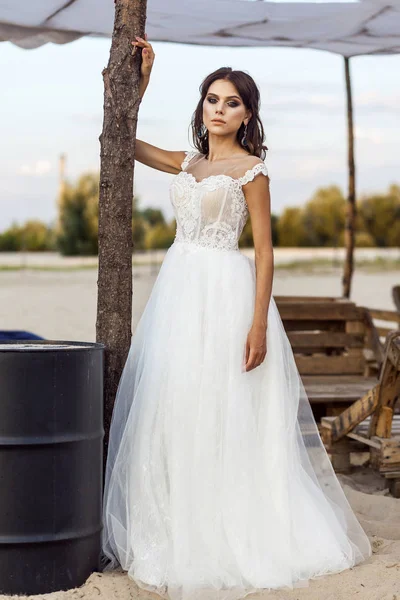 attractive brunette woman with makeup and hairstyle wearing white wedding dress while posing outside