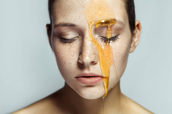 Portrait of beautiful young brunette woman with closed eyes and freckles with honey on face against gray background.