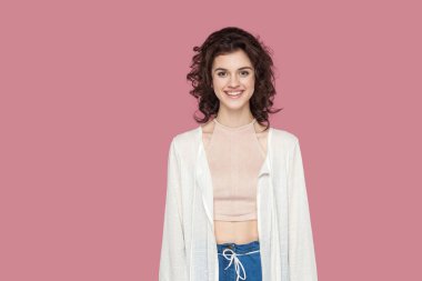 rahat tarzı ayakta kıvırcık saç modeli ile mutlu memnun güzel esmer genç kadın ayakta ve pembe arka plan üzerinde dişli gülümseme ile kamera bakıyor