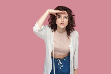 alnında el ile ayakta ve pembe arka plan üzerinde uzağa bakarak rahat tarzı kıvırcık saç modeli ile ciddi özenli güzel esmer genç kadın