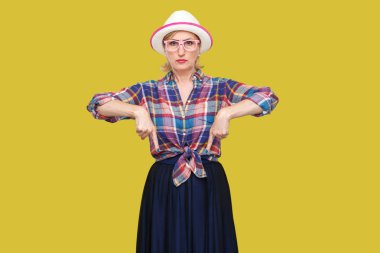 sarı arka plan üzerinde kamera bakarken ayakta ve aşağı işaret şapka ve gözlük ile rahat tarzı ciddi modern şık olgun kadın