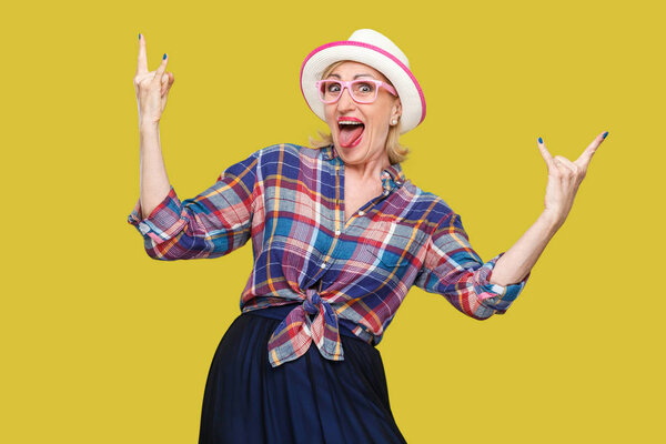сумасшедшая смешная современная стильная зрелая женщина в непринужденном стиле со шляпой и очками, показывающими рок-знаки и язык, глядя на камеру на желтом фоне
