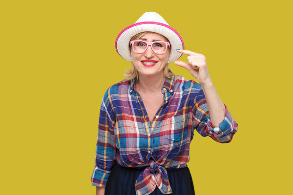 порадовала современная стильная зрелая женщина в непринужденном стиле с шляпой и очками стоя и показывая немного жеста с пальцами на желтом фоне
