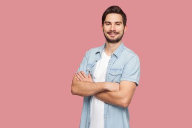 mavi rahat tarzı gömlek mutlu başarılı yakışıklı sakallı genç adam çapraz kolları ile ayakta ve pembe arka plan üzerinde dişli gülümseme ile kameraya bakıyor