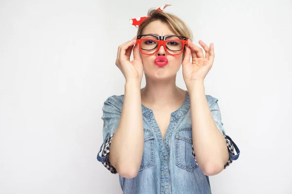 funny young woman in casual blue denim shirt with makeup and red headband wearing eyeglasses frames and sending air kiss at camera isolated on white background