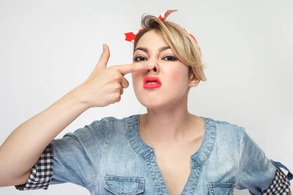 uncultured young woman in casual blue denim shirt with makeup and red headband pointing finger on nose while showing bad smell isolated on white background