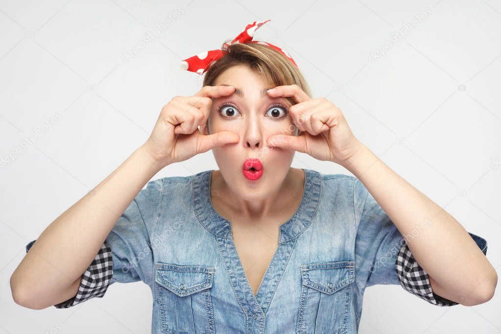 wondering beautiful young woman in casual blue denim shirt with makeup and red headband making eyeglasses with fingers and looking at camera with shocked face isolated on white background